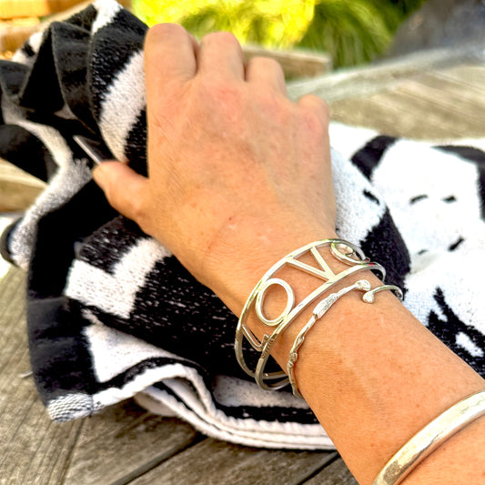 A close-up of a person's wrist wearing three silver-toned bracelets, including a cuff with the word 'LOVE' in openwork letters, on a black and white striped towel over a wooden table.